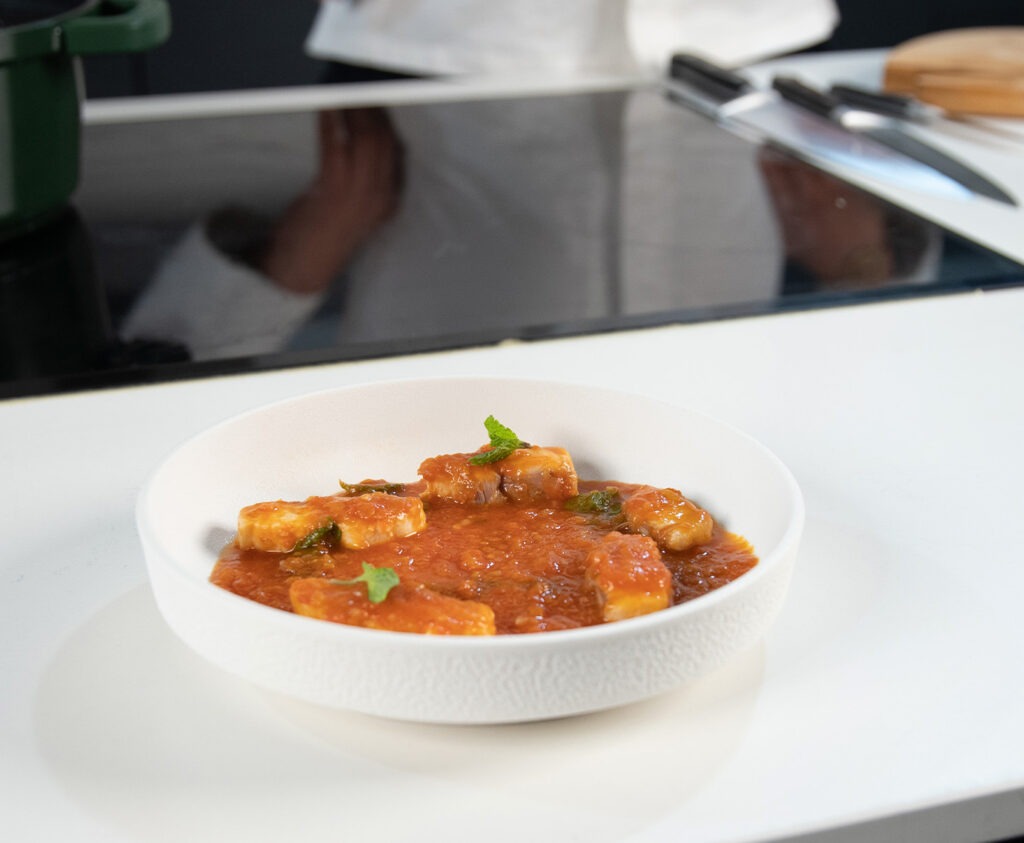 Cazón en tomate tradicional gaditano servido en plato hondo blanco con salsa de tomate espesa y pan al lado